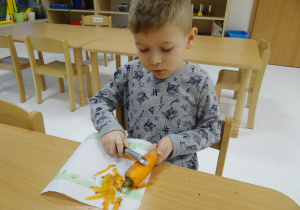 Szymonek siedzi przy stoliku i obiera marchewkę.